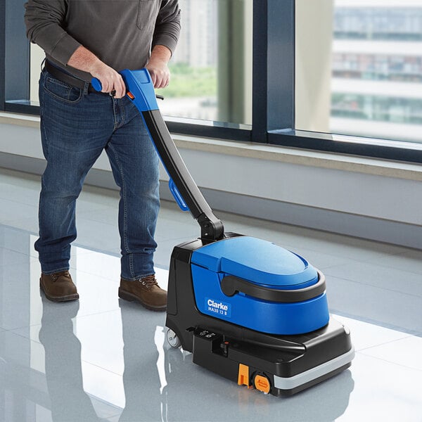 A person using a Clarke cordless walk behind cylindrical floor scrubber to clean a floor.
