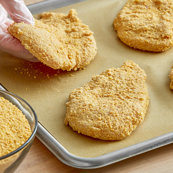 A gloved hand uses Kellogg's Corn Flake Crumbs to coat fried chicken on a baking sheet.