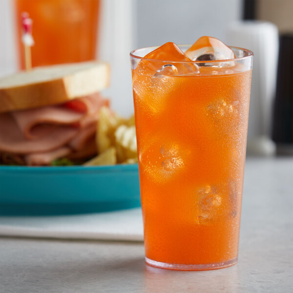 A close-up of a clear plastic tumbler filled with orange liquid and ice with a sandwich in the background.
