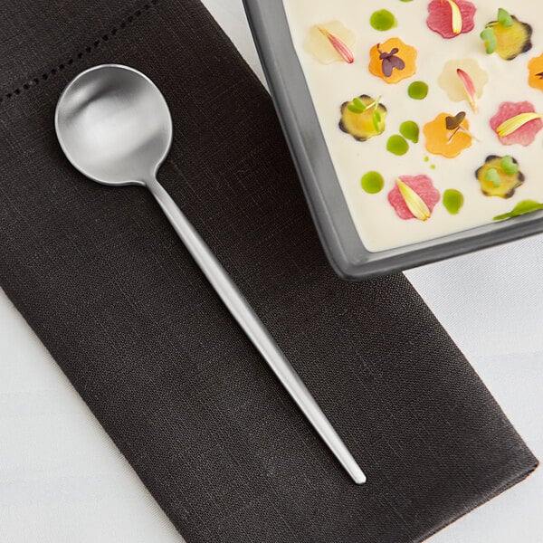 An Acopa brushed stainless steel bouillon spoon in a bowl of soup on a table.