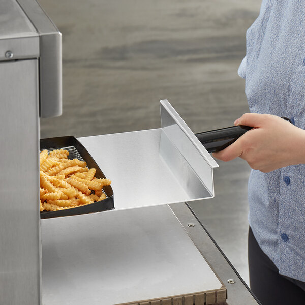 A woman using a 13 1/2" x 9 1/2" aluminum paddle peel with a black handle to remove french fries from an oven.
