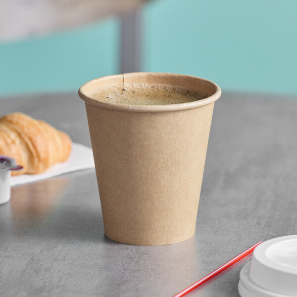 A brown Kraft paper hot cup filled with coffee on a saucer with a croissant.