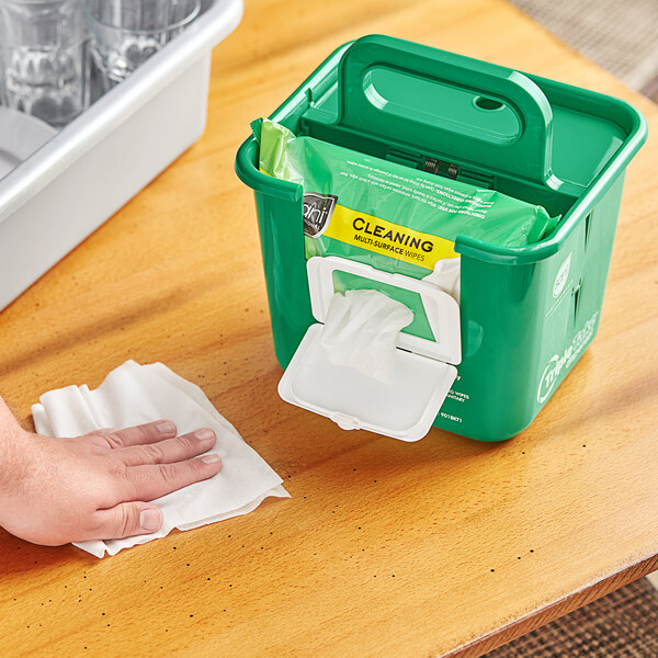 A hand wiping a kitchen counter with a white tissue from a green Sani Professional Triple Take dispenser.