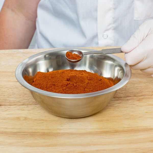 A person holding a spoon of red powder in a Cardinal Detecto stainless steel mixing bowl.