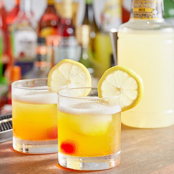 A bottle of Finest Call Premium Lemon Sour Mix on a table with two glasses of yellow liquid and lemon wedges.