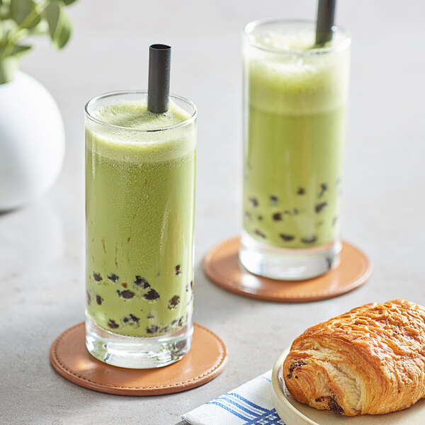 Two glasses of green matcha bubble tea with black straws and a croissant on a table.