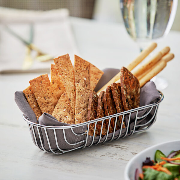 An Acopa oval chrome wire basket filled with crackers and bread sticks on a table.
