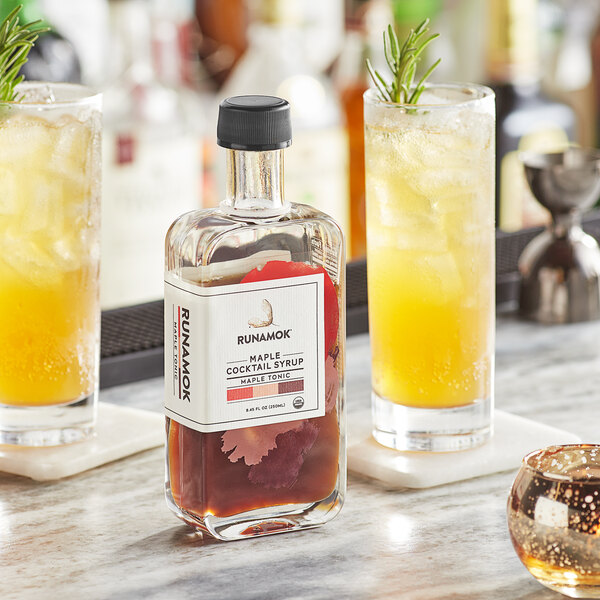 A bottle of Runamok Maple Tonic Cocktail Syrup on a bar counter next to a glass of tonic.