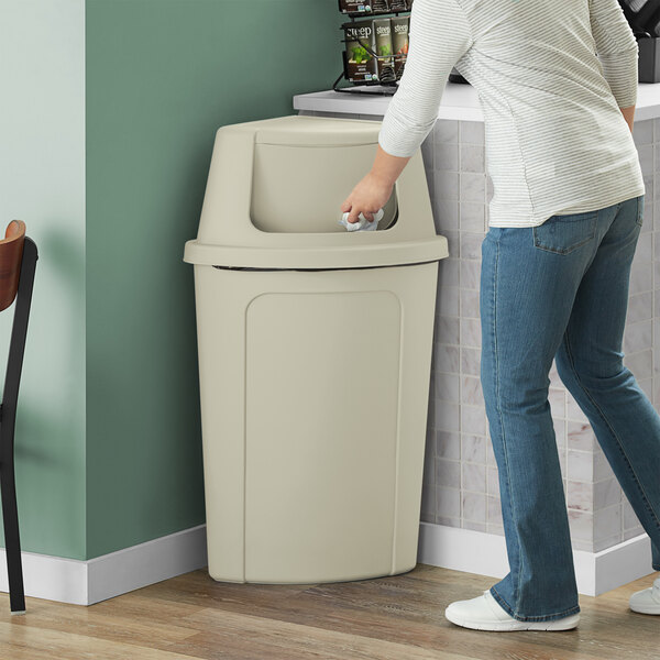 A woman using a Lavex beige corner round trash can with a beige push door lid to throw away paper.