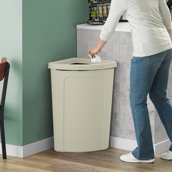 A woman putting a bag in a Lavex beige corner round trash can with a beige lid.