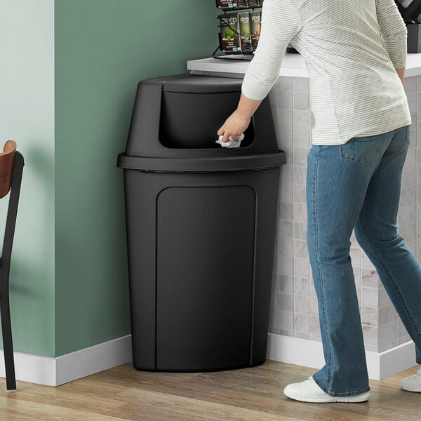 A woman in a white shirt and jeans opens a Lavex black corner round trash can with a black push door lid to throw away paper.