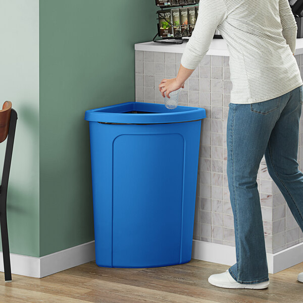 A woman standing next to a Lavex blue corner round trash can with a blue rim opening it to throw away a plastic cup.
