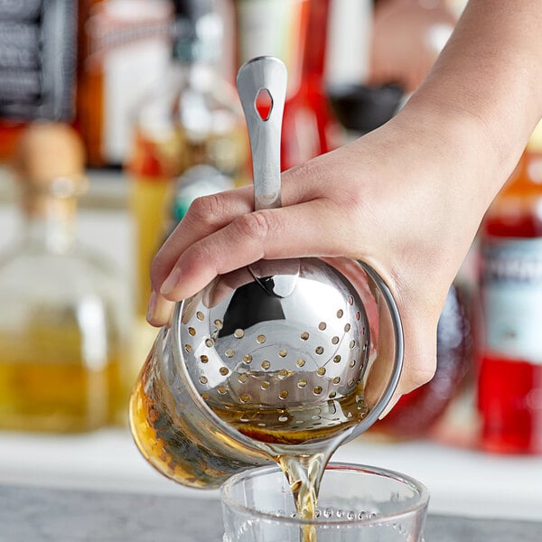 A hand using a Choice stainless steel Julep strainer to pour liquid into a glass on a counter.