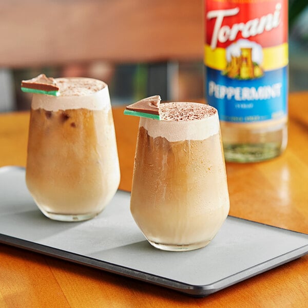 Two glasses of peppermint-flavored drinks on a tray with a bottle of Torani Peppermint syrup.