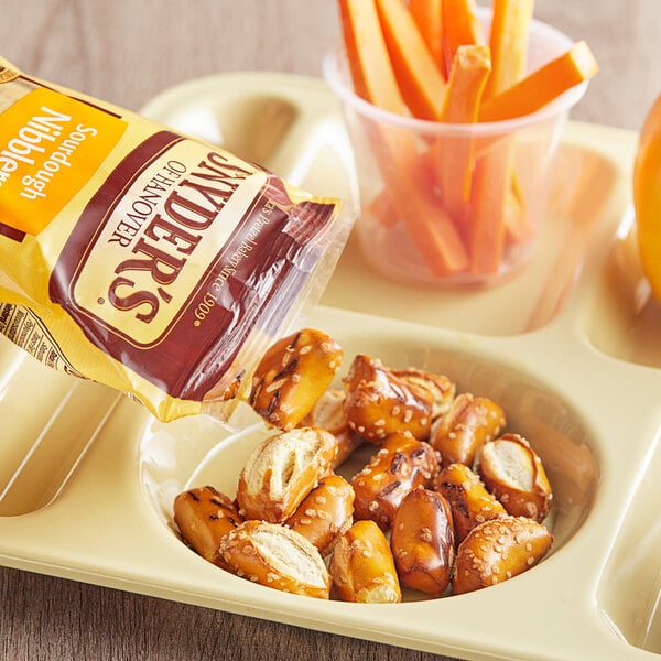A tray with a bag of Snyder's of Hanover Sourdough Pretzel Nibblers and a plastic cup of carrot sticks on a table.