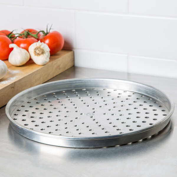 An American Metalcraft heavy weight aluminum pizza pan with perforated holes, with tomatoes and garlic on it.