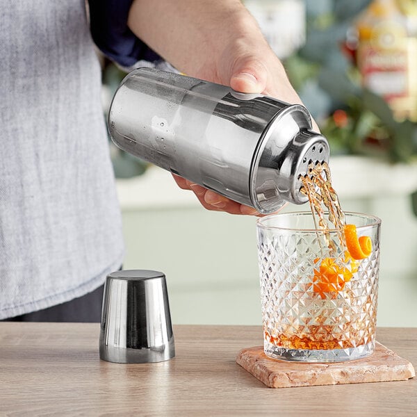 A hand using an Acopa stainless steel cocktail shaker to pour orange juice into a glass.