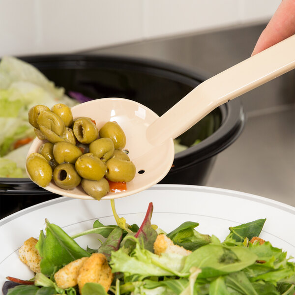 A person using a Thunder Group beige polycarbonate perforated salad bar spoon to serve olives from a bowl of salad.