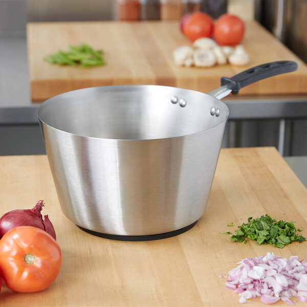 A Vollrath stainless steel sauce pan on a wood surface with vegetables.