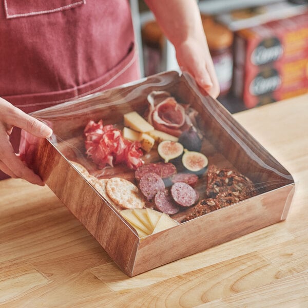 A person holding an Enjay wood laminated box filled with meat and cheese on a table in a deli.