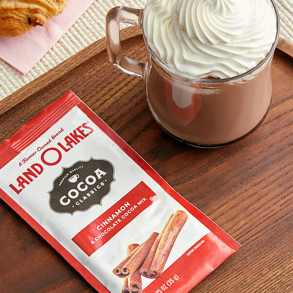 A glass mug of Land O Lakes hot chocolate with whipped cream and cinnamon sticks on a wooden tray.