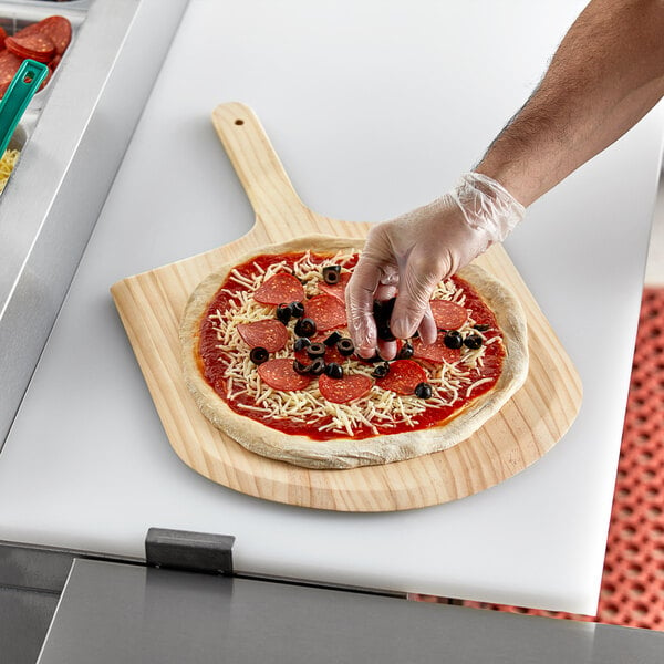 A person putting toppings on a pizza using a Choice wooden pizza peel.