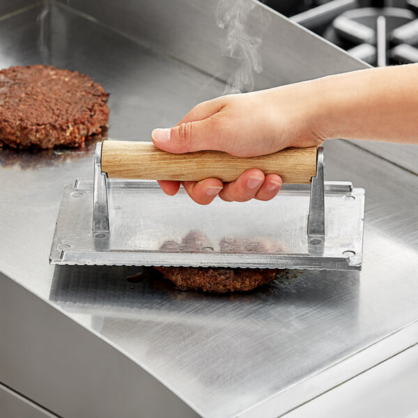 A person using a wooden-handled aluminum steak weight to grill a burger patty.