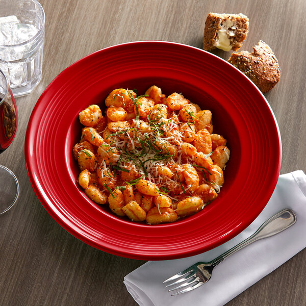 A red Tuxton Concentrix bowl filled with pasta and herbs on a table with a fork and glass of wine.