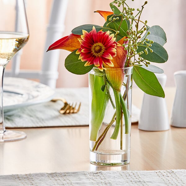 An Acopa cylindrical glass vase with flowers and leaves on a table.