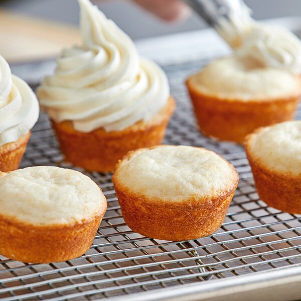 A person using a pastry brush to frost a group of Gold Medal white cupcakes on a cooling rack.