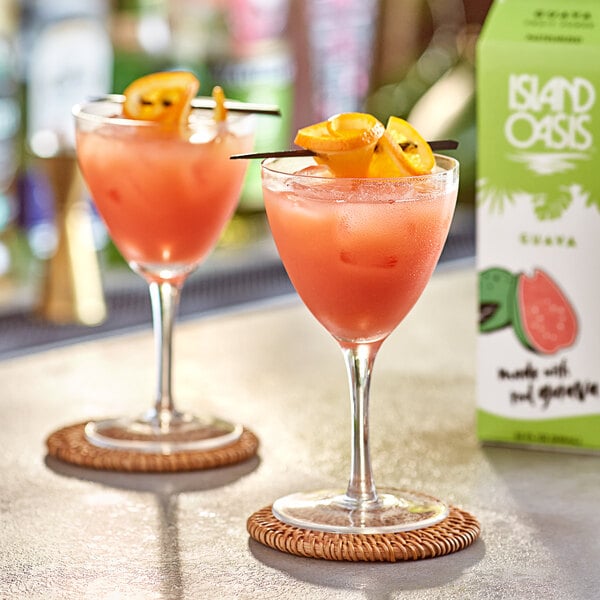 Two glasses of pink Island Oasis Guava drinks with orange slices and straws on a table.