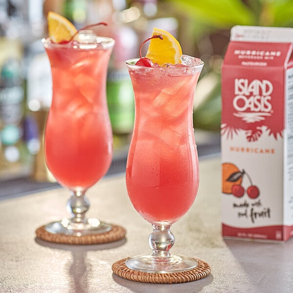 Two glasses of pink Island Oasis Hurricane drinks garnished with orange slices on a table.