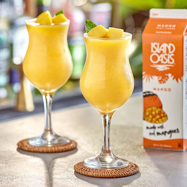 A table with two glasses of mango drinks, one garnished with orange, and a box of Island Oasis Mango Frozen Beverage Mix.