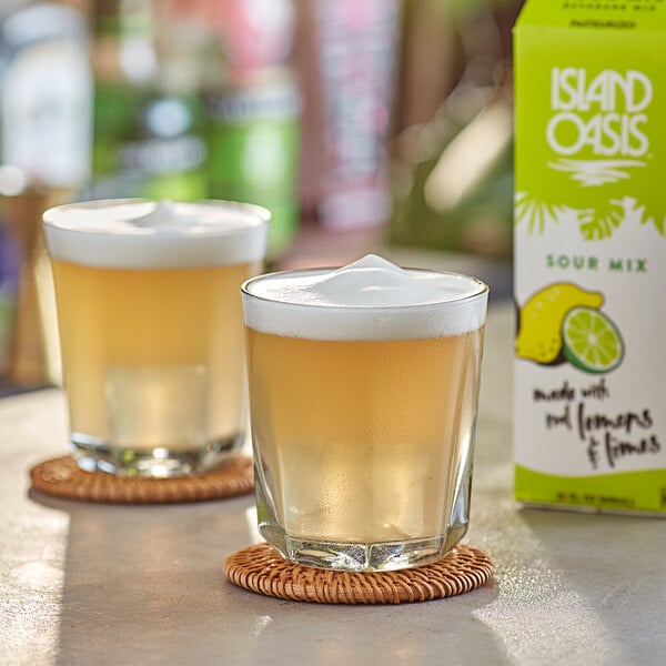 Two glasses of liquid on a coaster next to a box of Island Oasis Sour Frozen Beverage Mix.