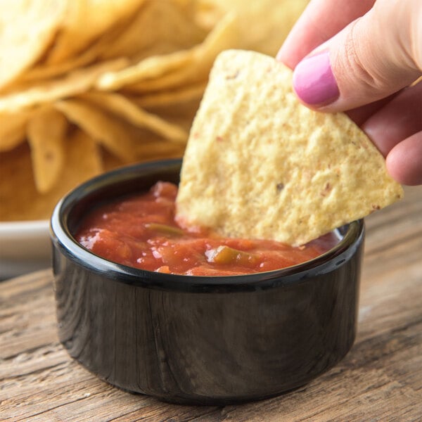 A person holding a chip dipping it into a black Carlisle ramekin of salsa.