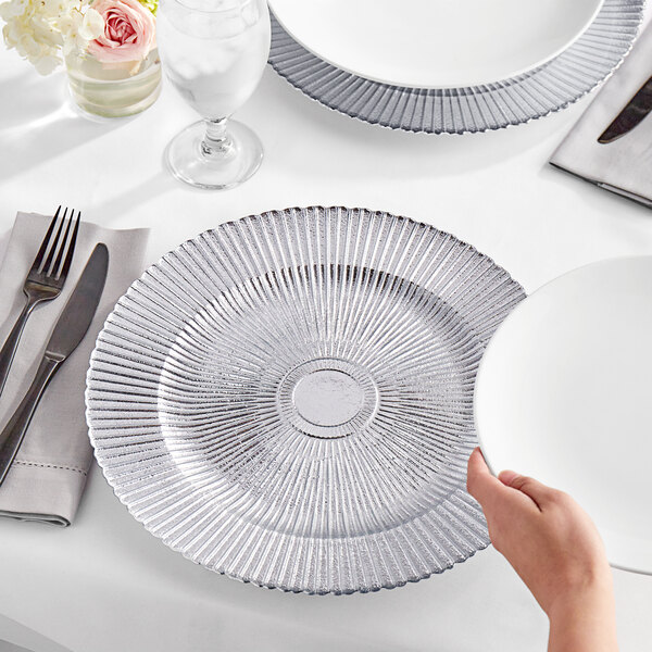 A hand holding an Acopa round silver sunburst glass charger plate on a table with a knife and a glass of water.
