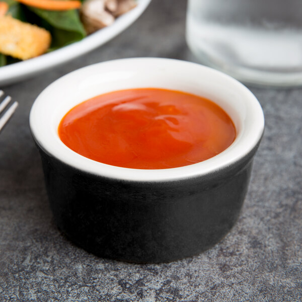 A Tuxton black and eggshell ramekin filled with red sauce on a table.