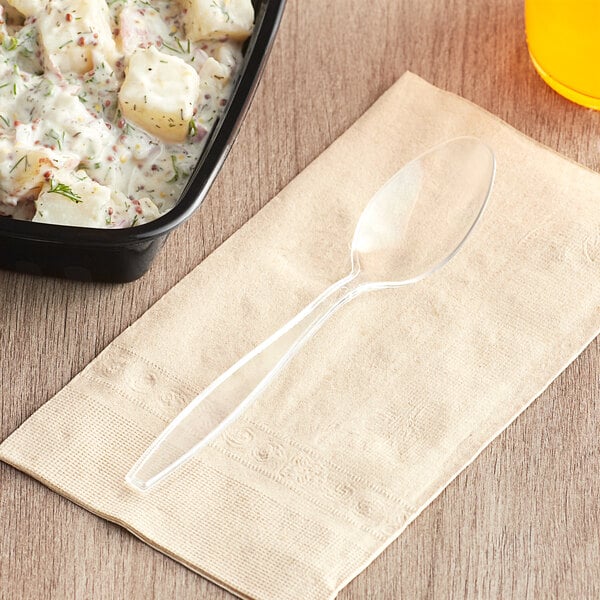 A Visions clear heavy weight plastic teaspoon on a napkin next to a bowl of food.