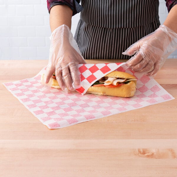 A person wearing gloves wrapping a sandwich in red check deli wrap paper.