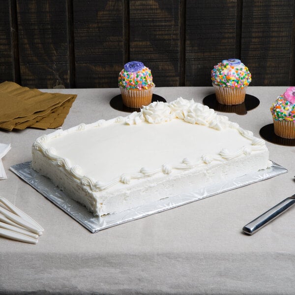 A white cake with frosting and cupcakes on a Enjay silver cake board on a table.