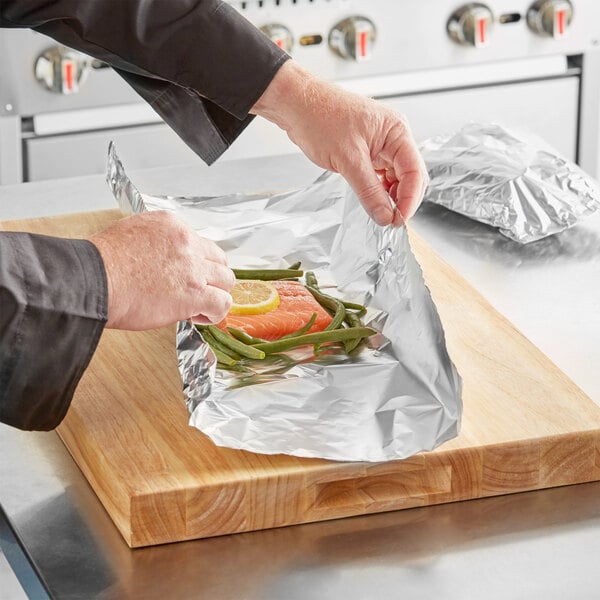 A person using Choice heavy-duty aluminum foil to wrap salmon with a lemon on a cutting board.
