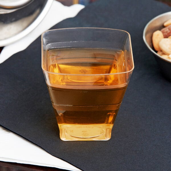 A Fineline clear plastic shot glass filled with brown liquid on a table with a bowl of nuts.