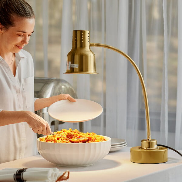 A woman using an Avantco gold heat lamp to keep food warm in a bowl on a table.