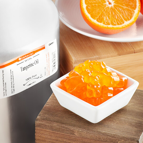 A close-up of a bottle of LorAnn Oils Tangerine Flavor next to a bowl of gummy bears and oranges.
