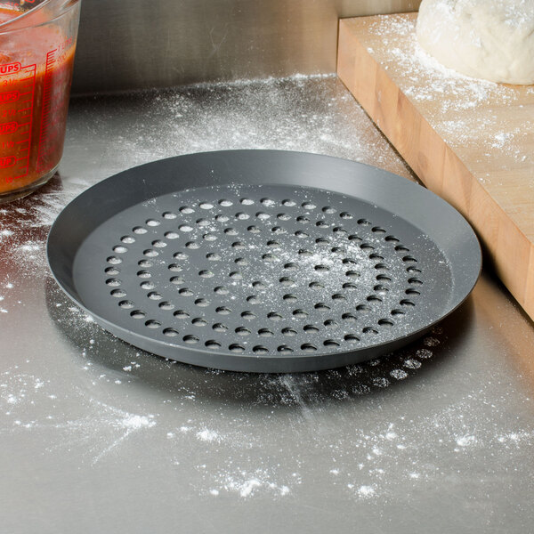 An American Metalcraft round black hard coat anodized aluminum pizza pan with holes on a counter with flour.