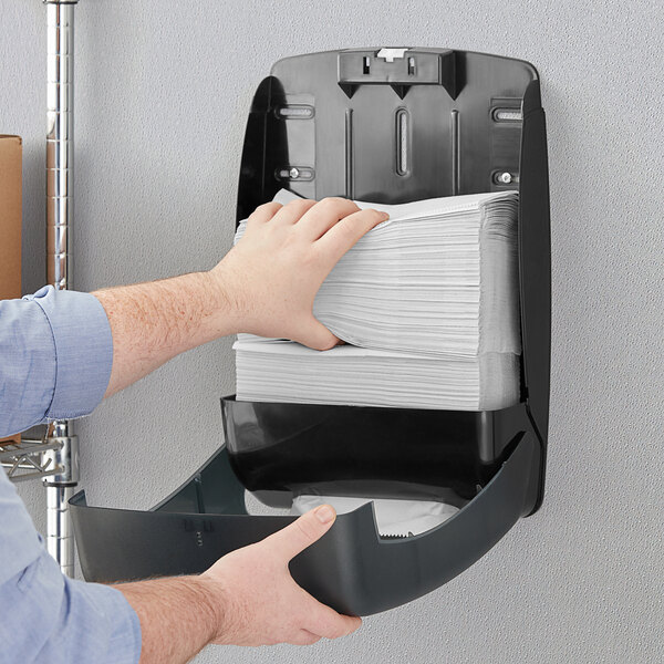 A hand putting Tork Universal white multi-fold paper towels in a wall mounted dispenser.