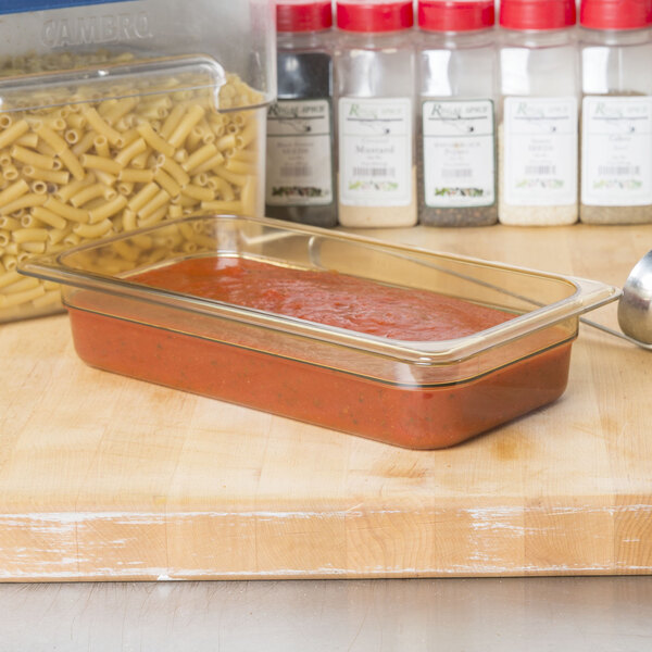 A Carlisle amber plastic food pan with pasta and sauce in it next to a container of pasta and a glass dish with sauce.