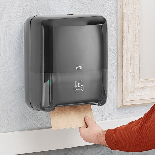 A person using a Tork black hands-free paper towel dispenser to put a paper towel in.