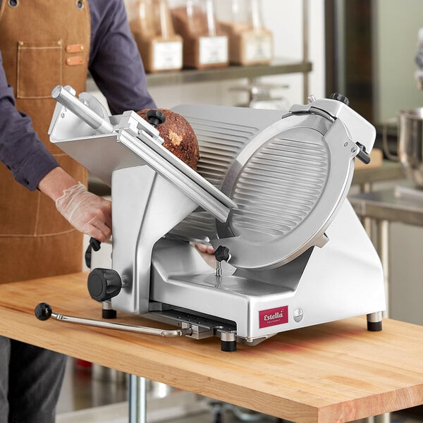 A person in an apron using a Estella manual meat slicer to cut meat on a table.