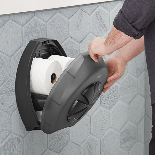 A person holding a Tork black three roll toilet paper dispenser in front of a wall.